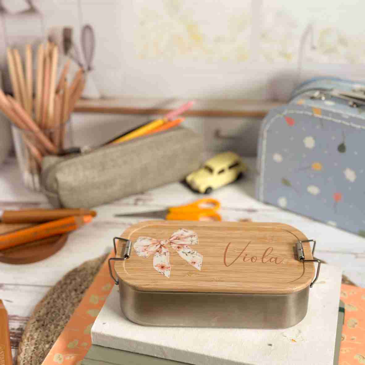 Personalisierte Edelstahl-Brotdose mit Bambusdeckel, floralem Schleifen-Motiv und dem Wunschnamen „Viola“.