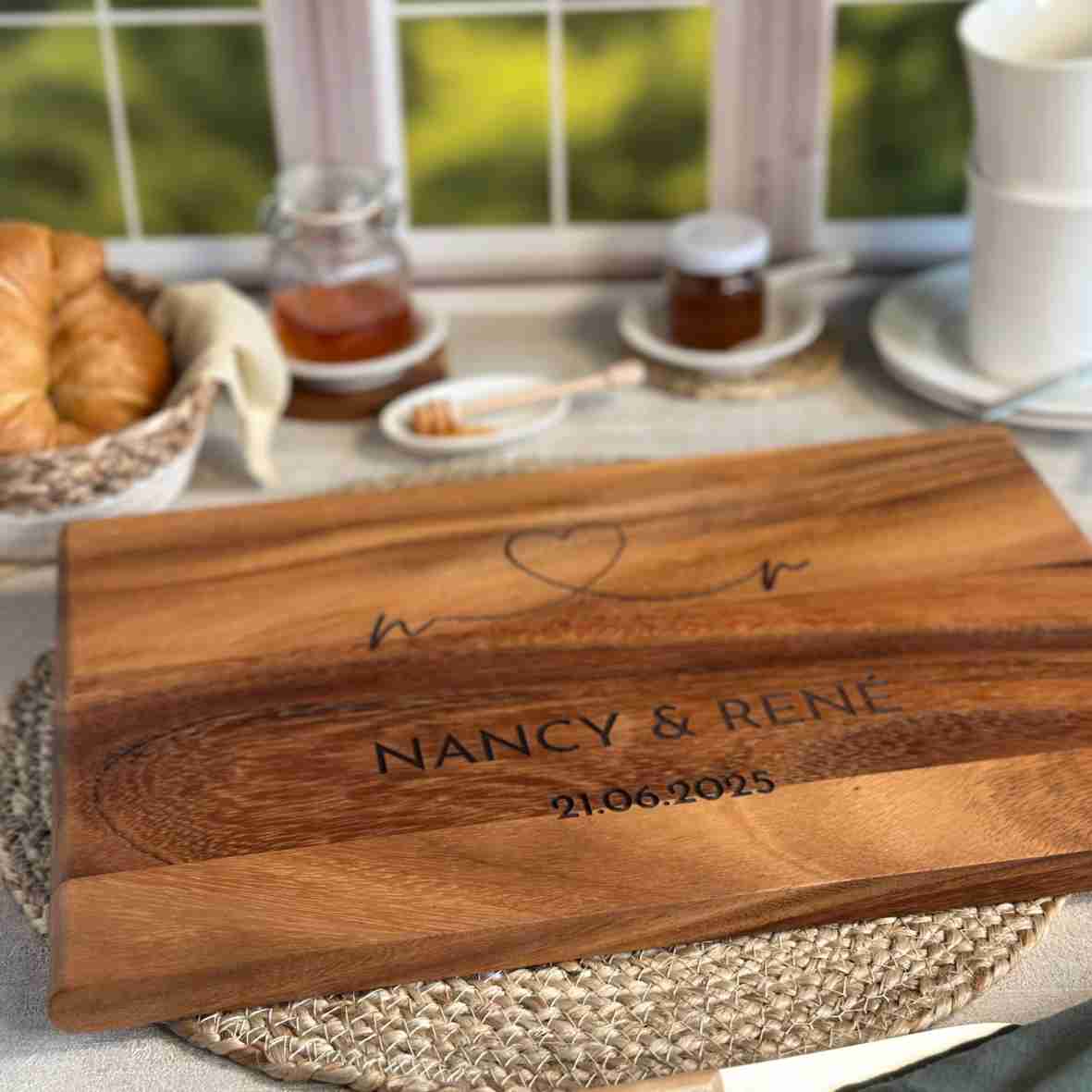 Personalisiertes Akazienholz-Schneidebrett mit Herzlinie, Gravur „Nancy & René“ und Datum 21.06.2025 in Frühstücksszene