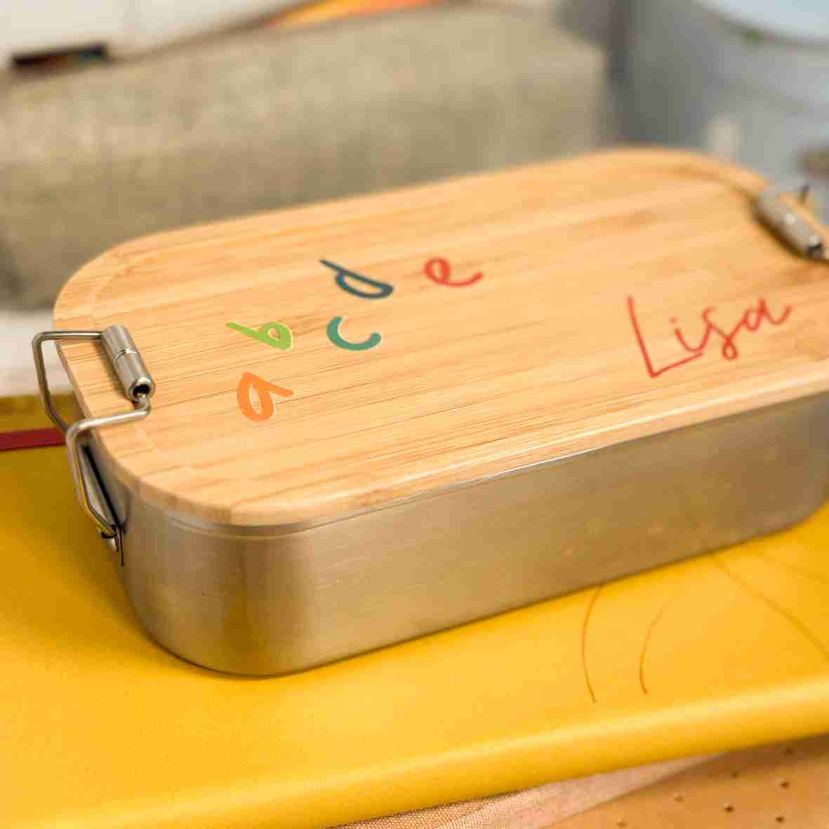 Personalisierte Brotdose aus Edelstahl mit Bambusdeckel, bedruckt mit bunten ABC-Buchstaben und dem Namen „Lisa“, auf einem Schulheft liegend, umgeben von Schulutensilien.