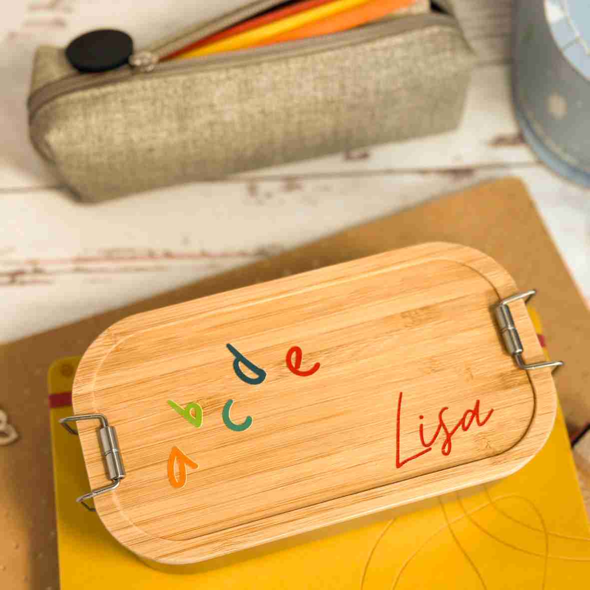 Personalisierte Brotdose aus Edelstahl mit Bambusdeckel, bedruckt mit bunten ABC-Buchstaben und dem Namen „Lisa“, auf einem Schulheft liegend, umgeben von Schulutensilien.