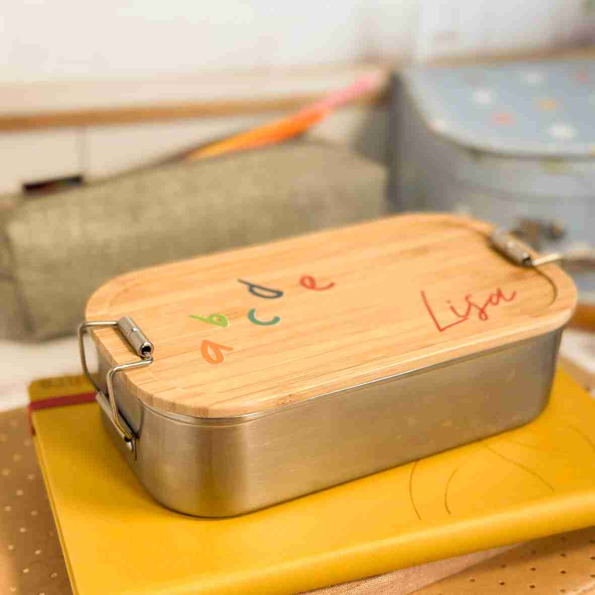 Personalisierte Brotdose aus Edelstahl mit Bambusdeckel, bedruckt mit bunten ABC-Buchstaben und dem Namen „Lisa“, auf einem Schulheft liegend, umgeben von Schulutensilien.