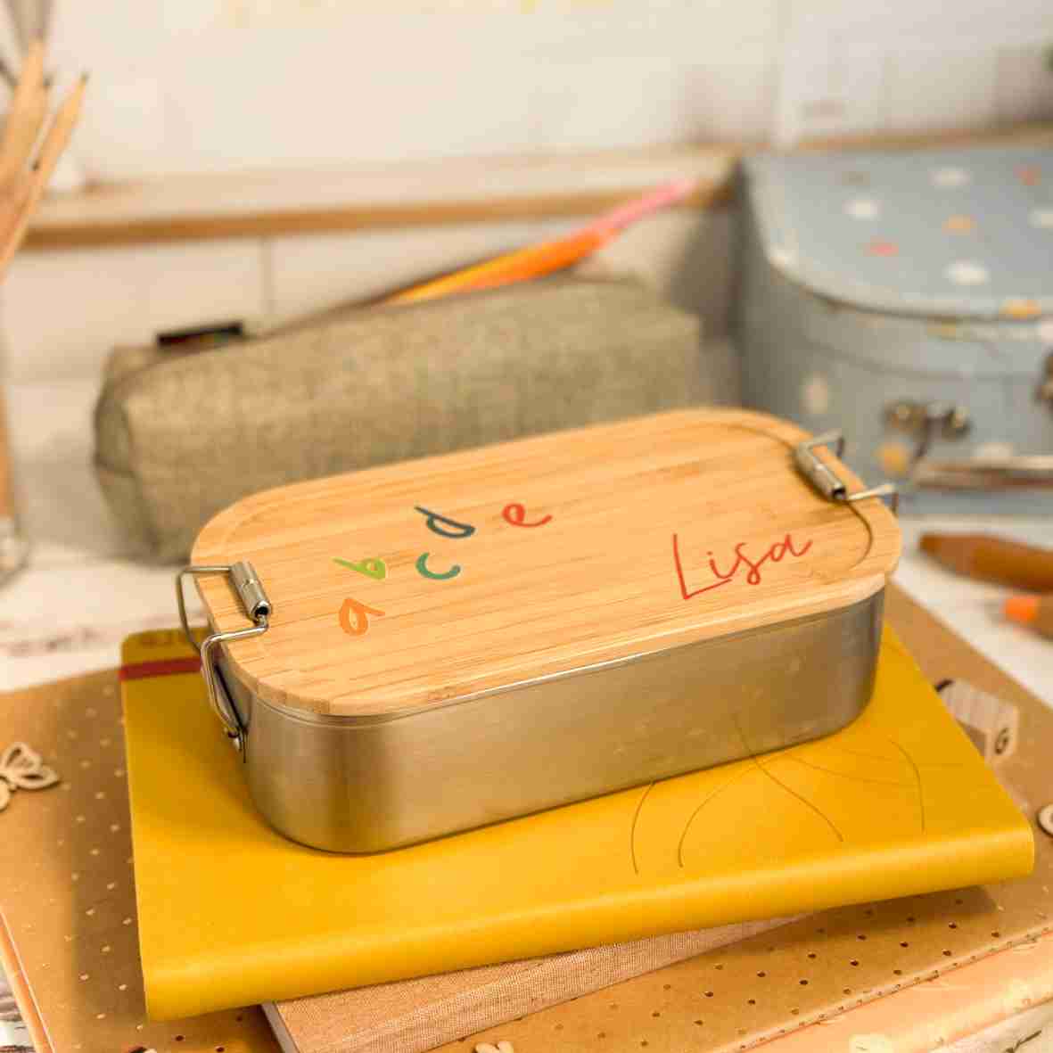 Personalisierte Brotdose aus Edelstahl mit Bambusdeckel, bedruckt mit bunten ABC-Buchstaben und dem Namen „Lisa“, auf einem Schulheft liegend, umgeben von Schulutensilien.