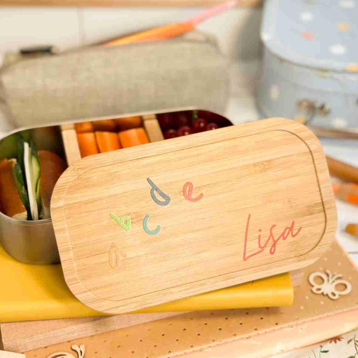 Personalisierte Brotdose aus Edelstahl mit Bambusdeckel, bedruckt mit bunten ABC-Buchstaben und dem Namen „Lisa“, auf einem Schulheft liegend, umgeben von Schulutensilien.