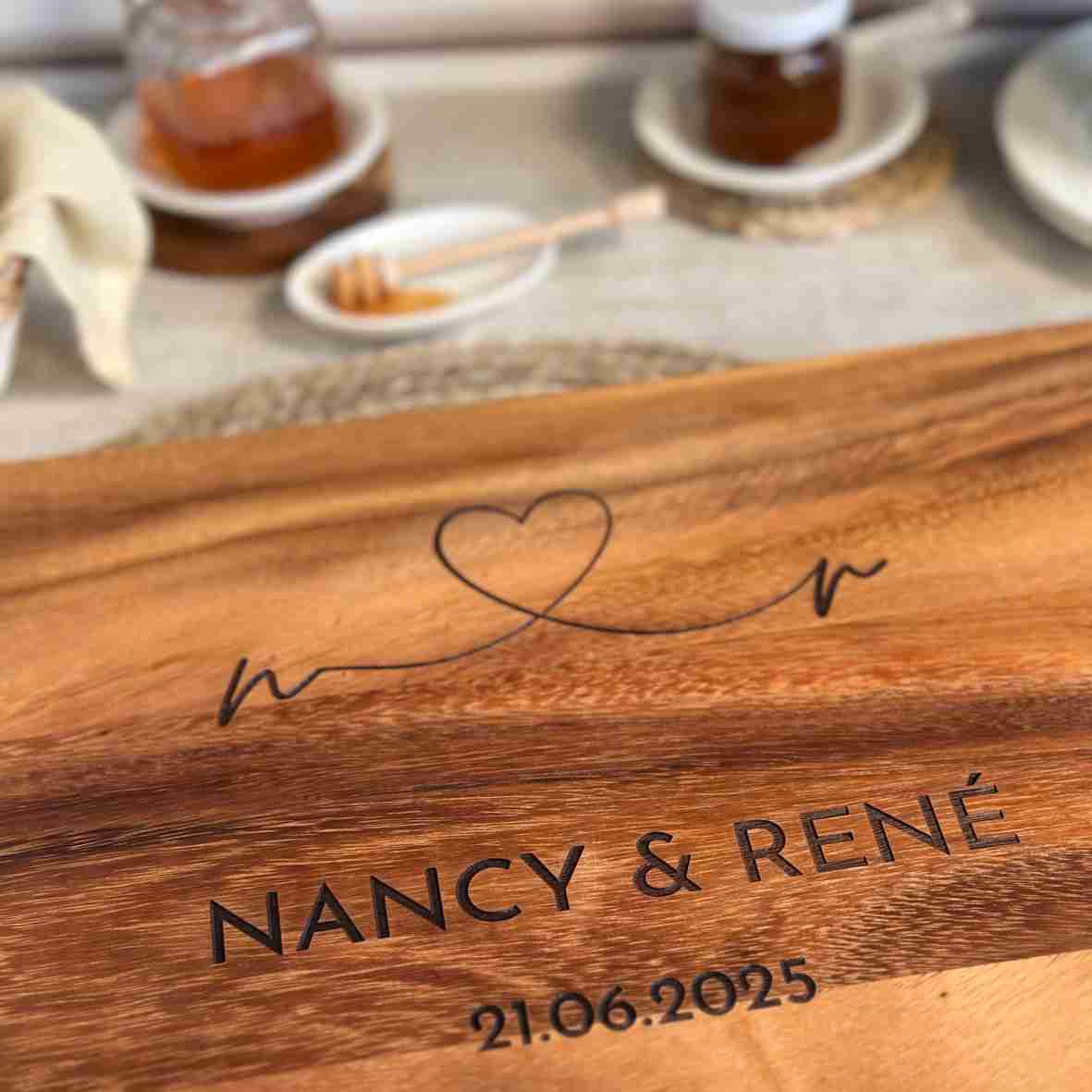 Personalisiertes Akazienholz-Schneidebrett mit Herzlinie, Gravur „Nancy & René“ und Datum 21.06.2025 in Frühstücksszene