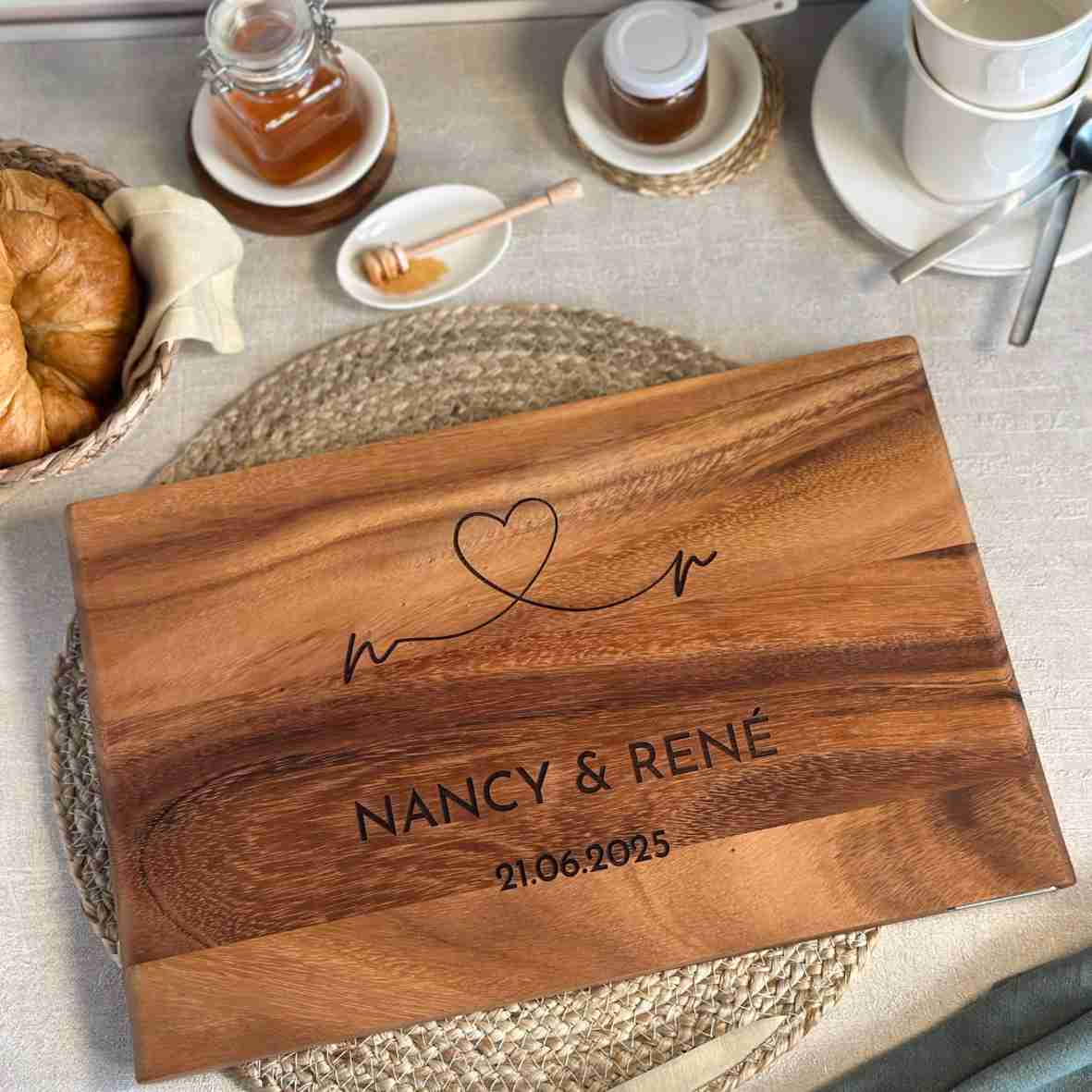 Personalisiertes Akazienholz-Schneidebrett mit Herzlinie, Gravur „Nancy & René“ und Datum 21.06.2025 in Frühstücksszene