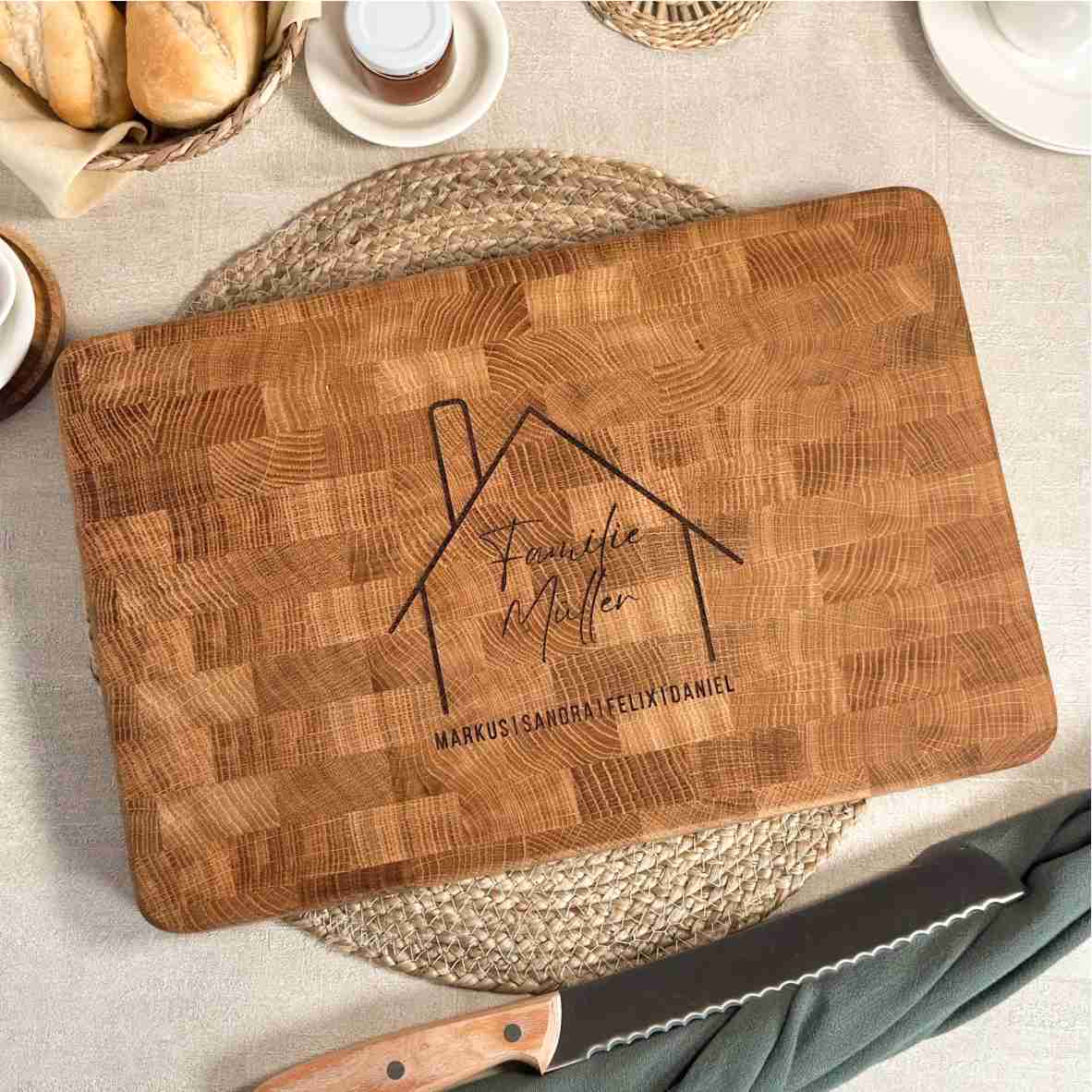 Personalisiertes Schneidebrett aus hochwertigem Eichen-Stirnholz, graviert mit stilisiertem Haus, Familiennamen und Vornamen, dekorativ präsentiert auf einem gedeckten Tisch. Ideal als Geschenk zum Einzug oder Hauskauf.