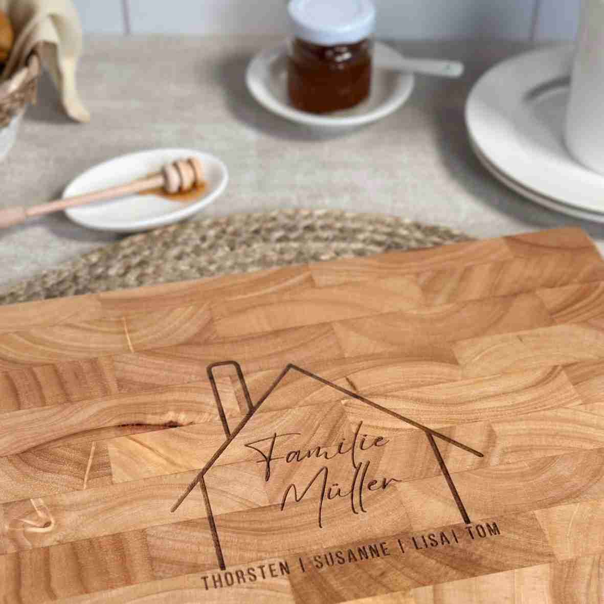 Graviertes Schneidebrett aus Stirnholz mit stilisierter Hausgrafik, Familiennamen „Familie Müller“ und den Vornamen aller Bewohner – Geschenkidee zum Einzug oder Hauskauf