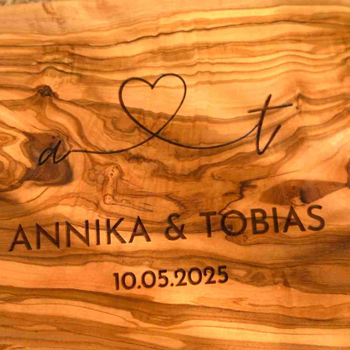 Personalisiertes Schneidebrett aus Olivenholz mit Herzgravur, Namen „Annika & Tobias“ und Datum „10.05.2025“, rustikal serviert auf einem gedeckten Tisch.