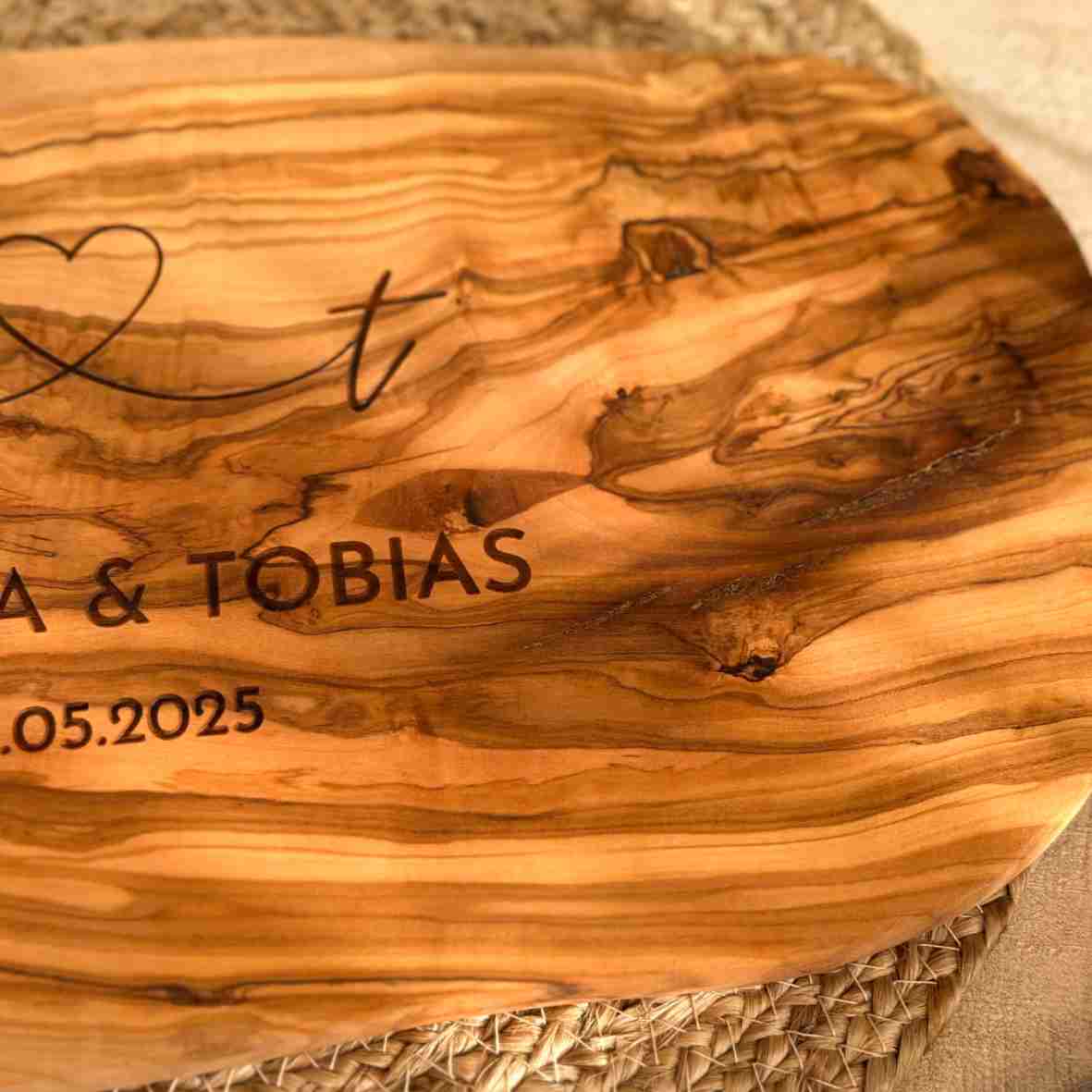 Personalisiertes Schneidebrett aus Olivenholz mit Herzgravur, Namen „Annika & Tobias“ und Datum „10.05.2025“, rustikal serviert auf einem gedeckten Tisch.