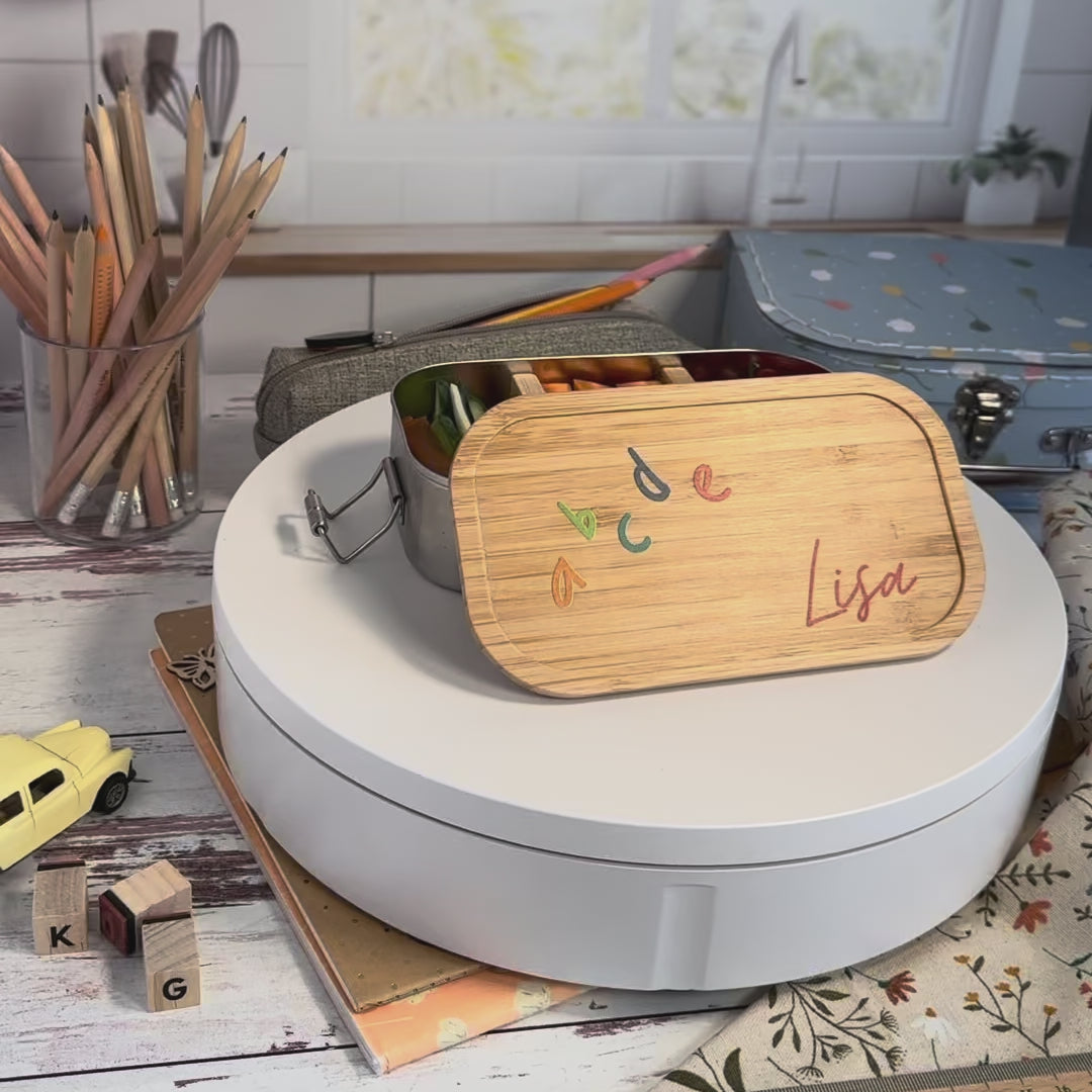 Personalisierte Brotdose aus Edelstahl mit Bambusdeckel, bedruckt mit bunten ABC-Buchstaben und dem Namen „Lisa“, auf einem Schulheft liegend, umgeben von Schulutensilien.
